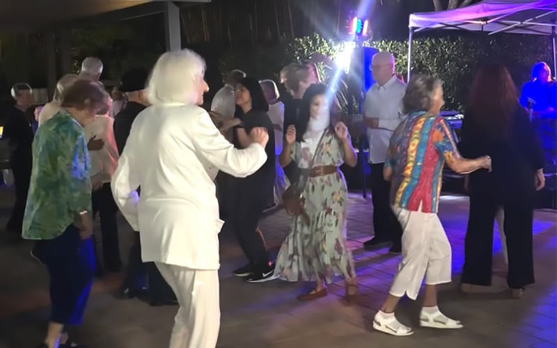 A group of people of various ages dance together outdoors at night under bright lights, some wearing colorful outfits. The background includes trees, a canopy, and a DJ booth.