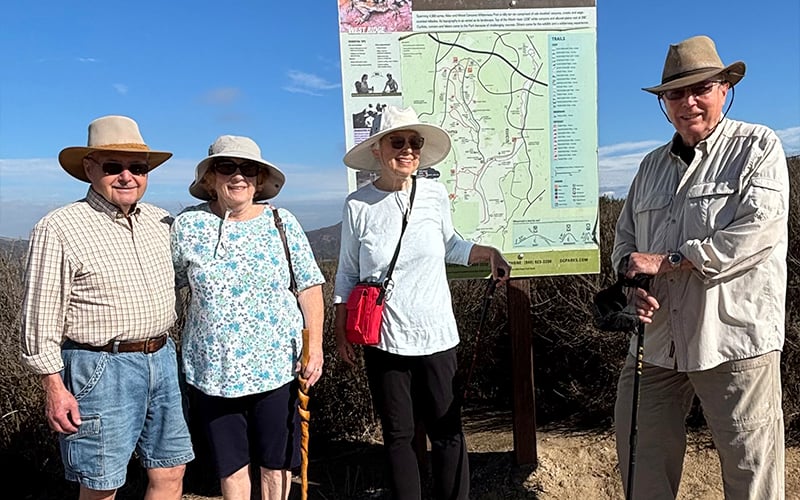 Four older adults wearing sun hats and sunglasses stand smiling in front of a large hiking trail map on a sunny day, dressed in light clothing and holding walking sticks. Dry brush and blue sky are visible in the background.