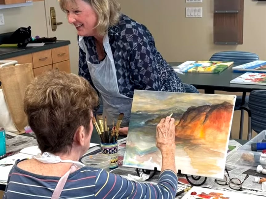Two women are at a table covered with art supplies and newspaper. One woman is painting a landscape on a canvas, while the other stands nearby, smiling and watching her work. The setting appears to be an art class or studio.