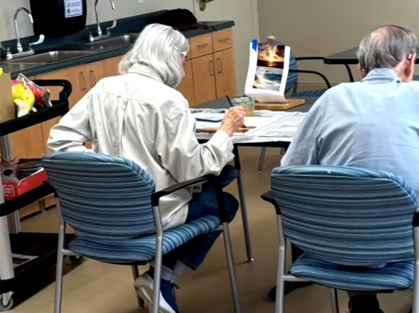 Two older adults sit at a table working on watercolor paintings, with art supplies spread out and a photograph of a sunset over water propped up as a reference. A cart with snacks is visible nearby.