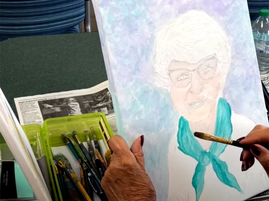 A person with painted nails holds a brush, working on a portrait of an elderly woman with glasses and a turquoise scarf. Paintbrushes and art supplies are arranged nearby on a table.