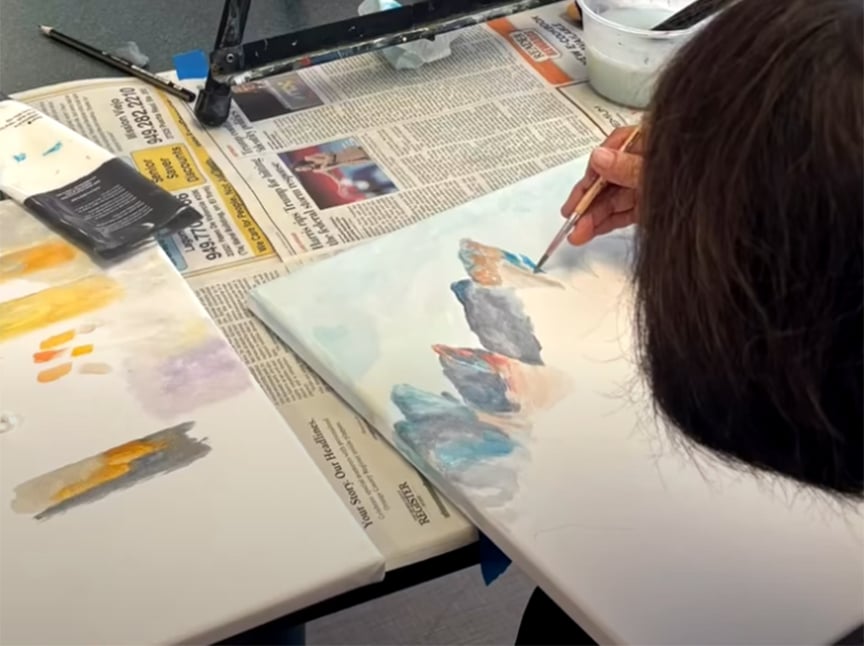 A person with dark hair paints colorful abstract shapes on a canvas, using a fine brush. Another canvas with color swatches and a newspaper are on the table beside them.