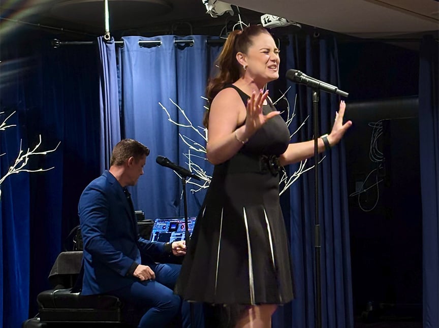 A woman in a black dress passionately sings into a microphone on stage, while a man in a blue suit plays piano in the background. The stage is decorated with blue curtains and white tree branches.
