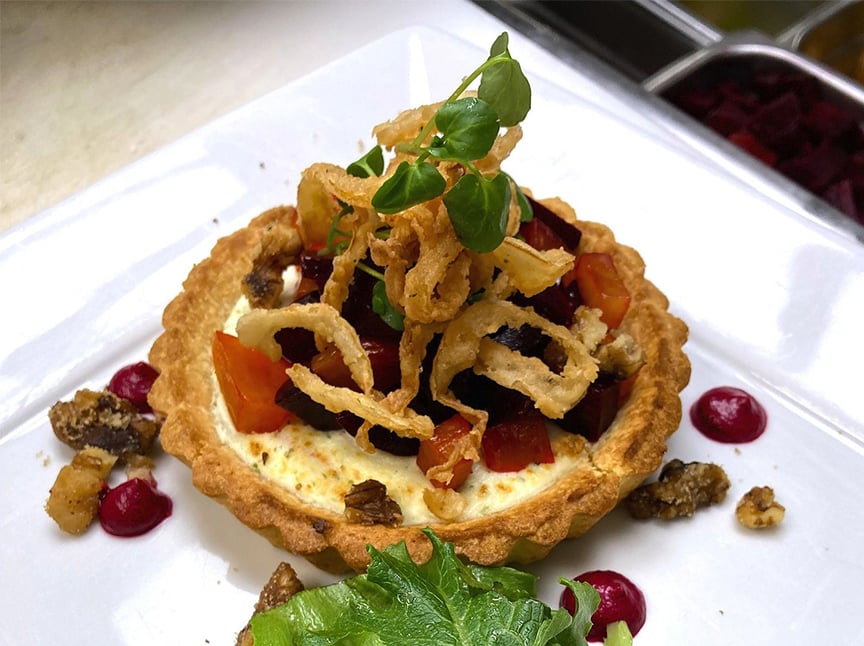 A savory tart topped with diced vegetables and crispy fried onions, garnished with microgreens. The plate is decorated with red sauce dots, walnuts, and a small leafy green salad.