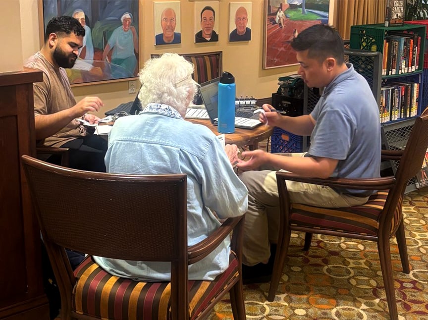 Two men sit at a table giving a manicure to an elderly woman with white hair in a cozy room with portraits on the wall and bookshelves nearby.
