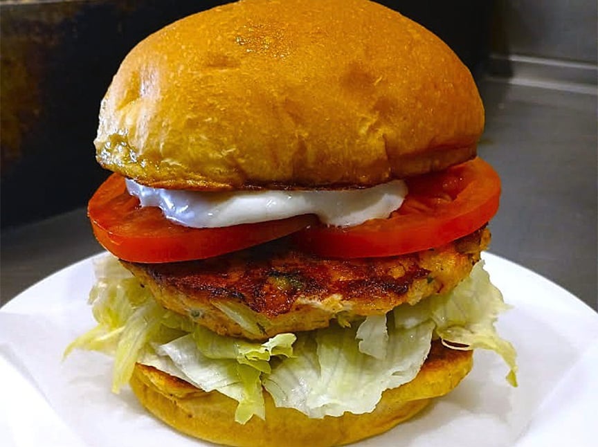 A close-up of a chicken burger with a golden bun, sliced tomatoes, lettuce, a grilled chicken patty, and a dollop of mayonnaise, served on a white plate.