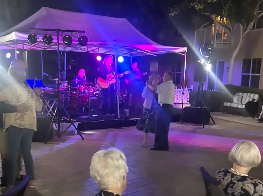 A live band performs under a tent with colorful stage lights as couples dance on an outdoor patio at night. People are seated in the foreground watching the performance.