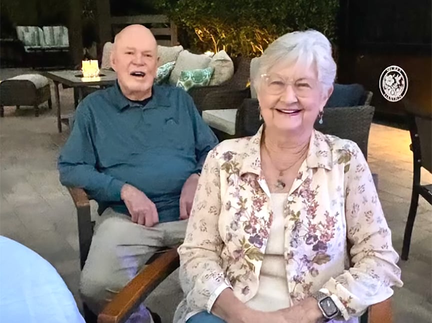 An elderly man and woman sit smiling on patio chairs outdoors in the evening, with cozy seating and candlelight in the background. The woman wears glasses and a floral shirt; the man wears a blue shirt.