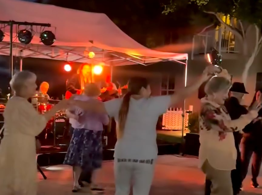 People of various ages dance together outdoors at night under string lights near a white tent, with a live band performing on stage in the background. The scene appears joyful and lively.