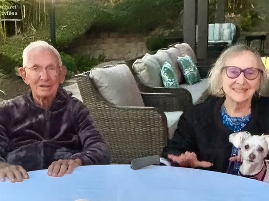 An elderly man and woman sit together at an outdoor table. The woman, wearing glasses, is holding a small white dog. Behind them are wicker sofas with patterned cushions and greenery in the background.