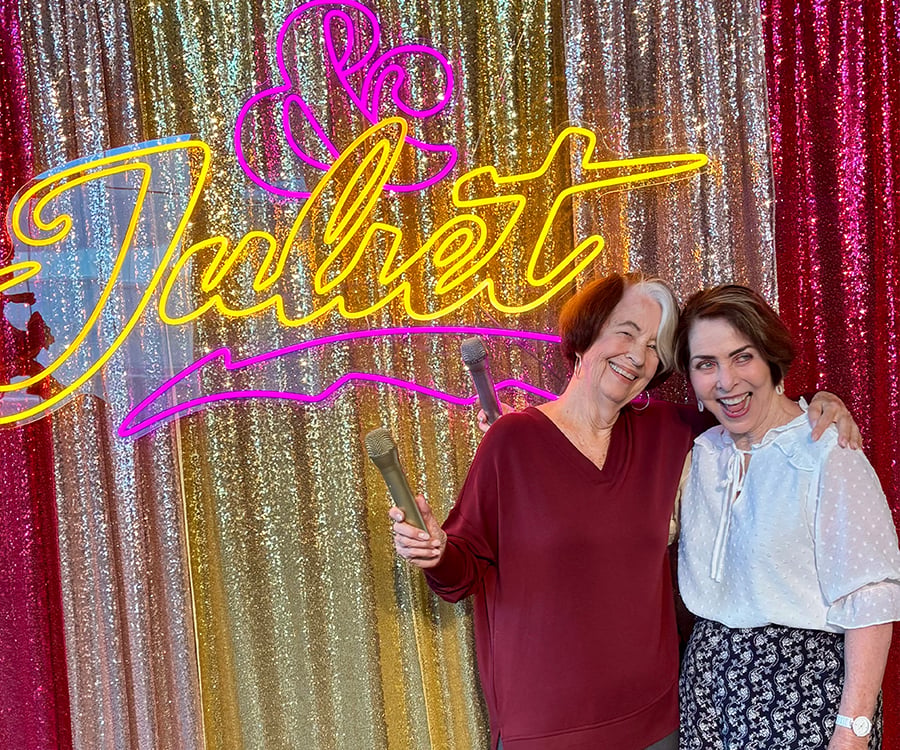 Two women smiling and holding microphones stand in front of a sparkling gold, pink, and red sequin backdrop with a bright neon sign that reads “& Juliet.”.