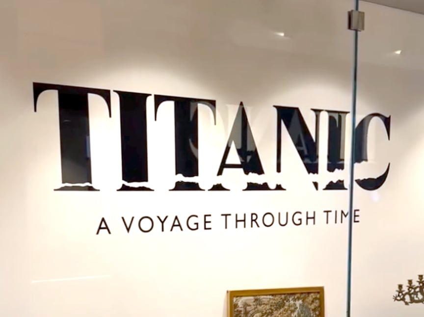 Large black text on a white wall reads TITANIC with smaller text underneath saying A VOYAGE THROUGH TIME. The image appears to be taken inside a museum or exhibition space.