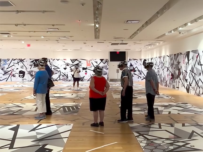 Several people wearing headsets stand in a modern art gallery with abstract black and white patterns covering the walls and floor, observing the space. The room is brightly lit with a wooden floor.