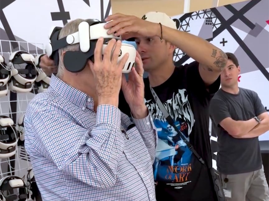 A young man helps an older man adjust a virtual reality headset. The older man is wearing a checkered shirt, and several VR headsets are visible in the background. Another person stands with arms crossed, watching.