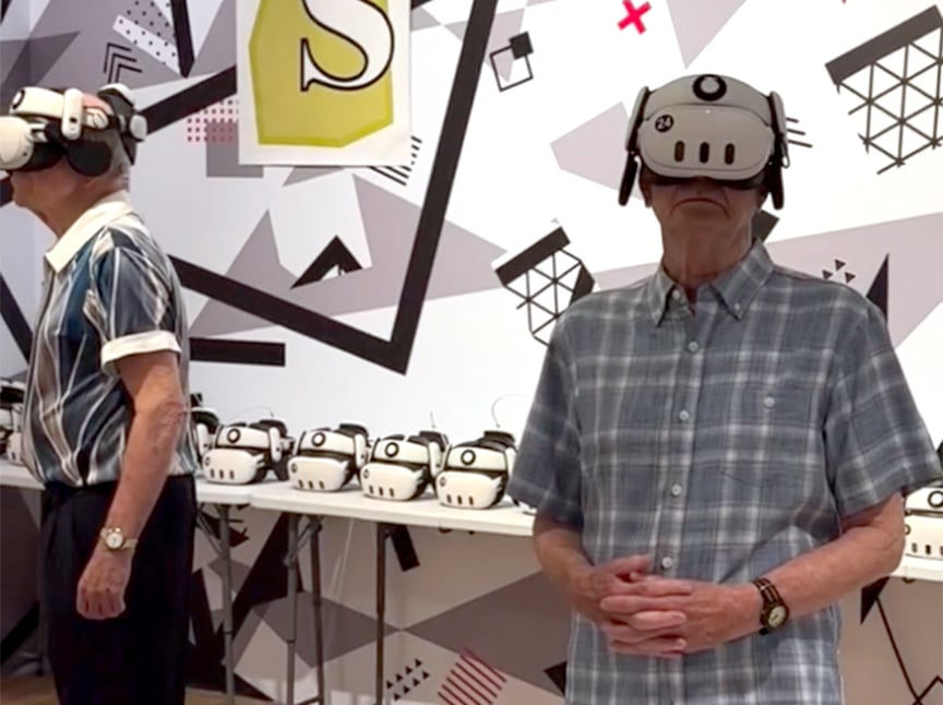Two older men wearing VR headsets stand in a modern, geometric-patterned room. A table behind them holds multiple similar VR headsets. Both men appear engaged with the virtual experience.