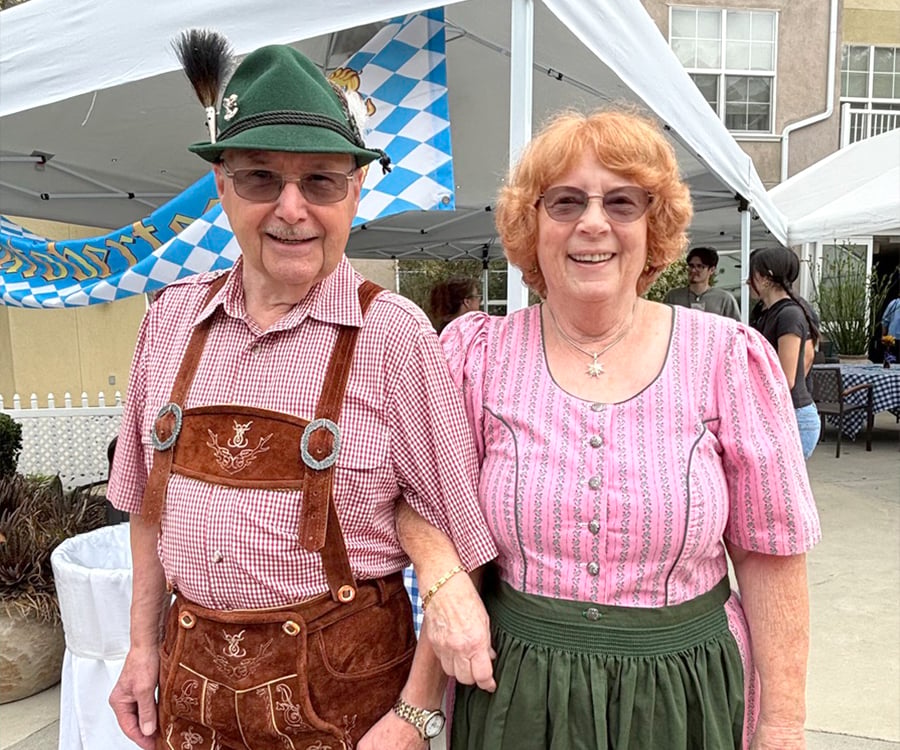 An older man and woman smile outdoors at a festival. The man wears lederhosen and a green hat with a feather, and the woman wears a traditional pink dirndl. Blue and white checkered banners hang in the background.