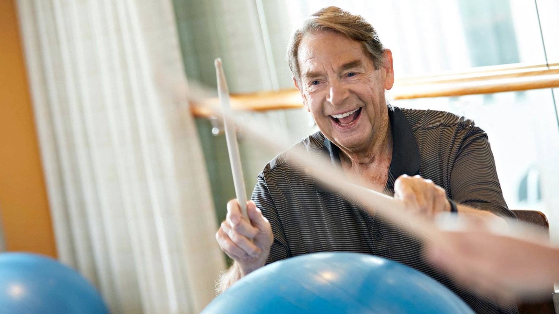An older man smiles and enthusiastically taps drumsticks on a large exercise ball during a fitness or therapy class indoors.