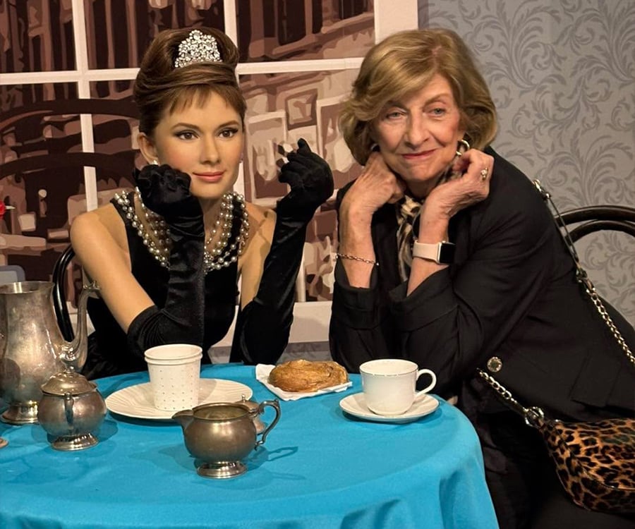 Two women pose at a table with tea and pastries; one is a wax figure dressed elegantly in black gloves and pearls, while the other is an older woman smiling and mimicking the pose.