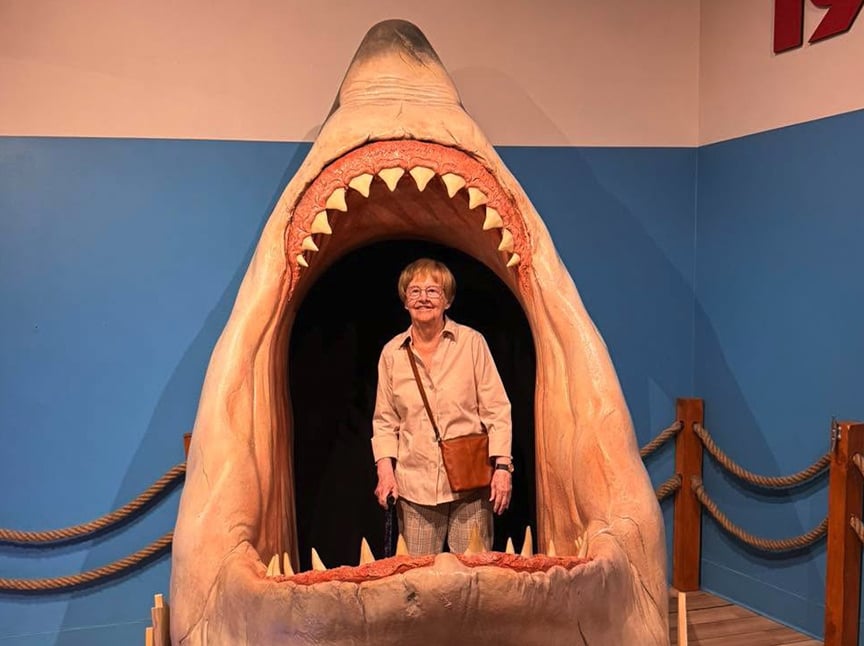 An older woman stands smiling inside a large model of an open shark’s mouth with big teeth, set against a blue and white wall with ropes on either side.