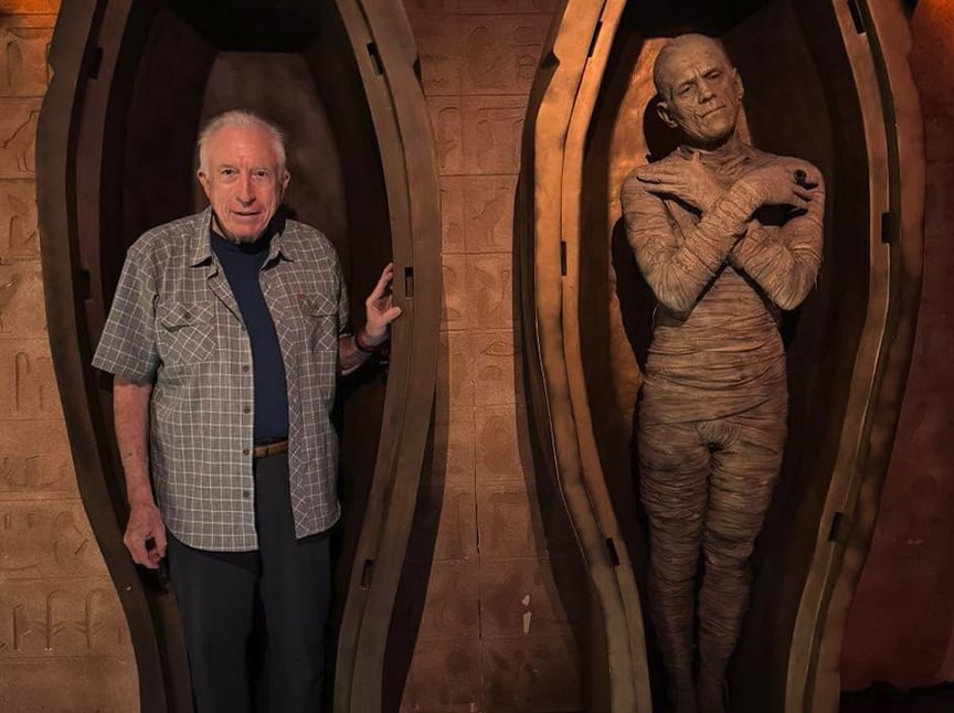 An elderly man stands inside an open sarcophagus next to a mummy figure wrapped in bandages, both set against a wall with Egyptian-style carvings.