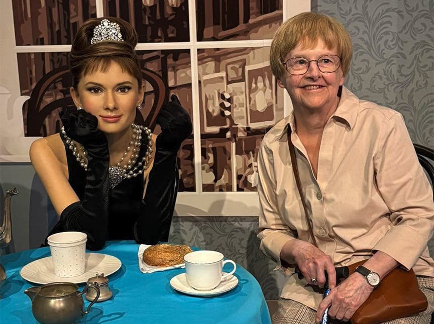 An older woman smiles while sitting next to a lifelike wax figure of Audrey Hepburn in a black dress and tiara, styled as in Breakfast at Tiffanys. They are seated at a table set with tea and pastries.