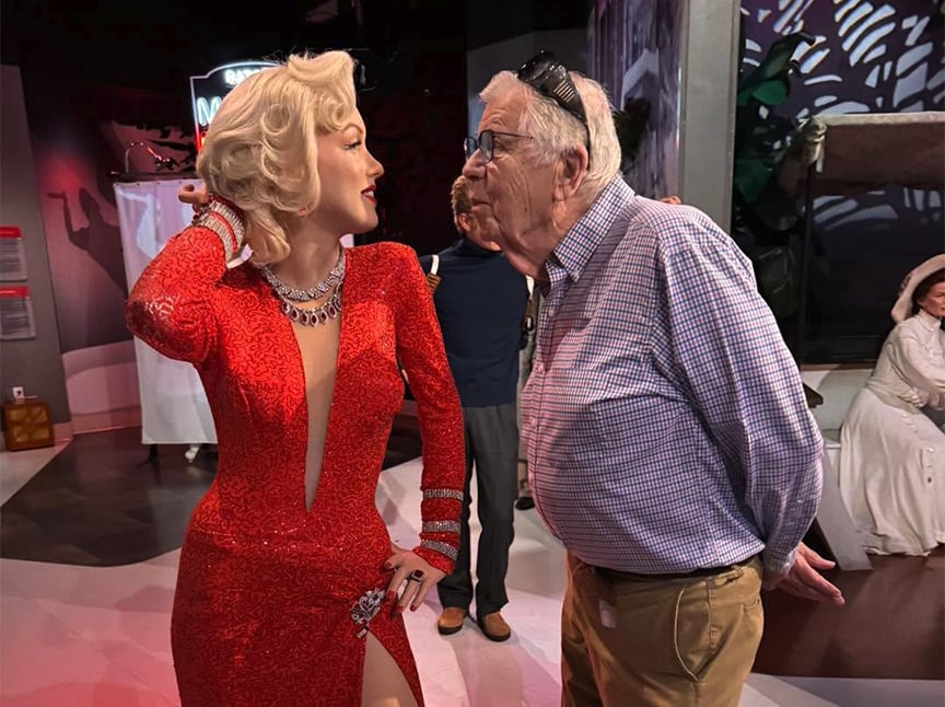 An older man leans in to kiss a lifelike wax figure of Marilyn Monroe dressed in a sparkling red gown at a museum or exhibit. Other figures and visitors are visible in the background.