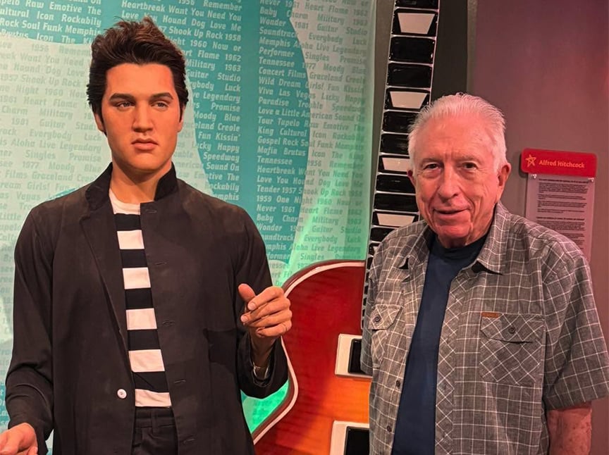 An older man stands next to a wax figure of Elvis Presley wearing a black jacket and striped shirt in a museum setting. A blue wall with song titles and a red guitar are in the background.
