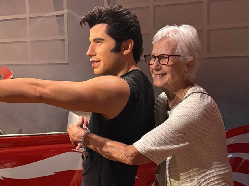 An elderly woman with glasses smiles and hugs a wax figure of a man in a black shirt who is pointing forward, standing next to a red and white car.