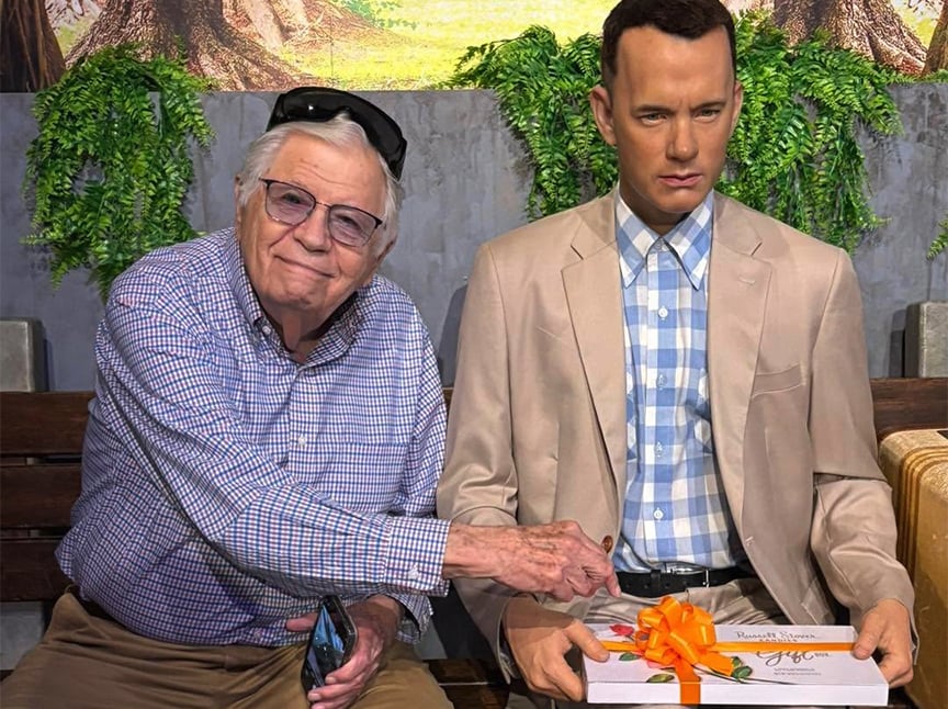 An elderly man in glasses smiles and poses with a lifelike wax figure of a man in a beige suit holding a box with an orange ribbon, both sitting on a bench with greenery in the background.