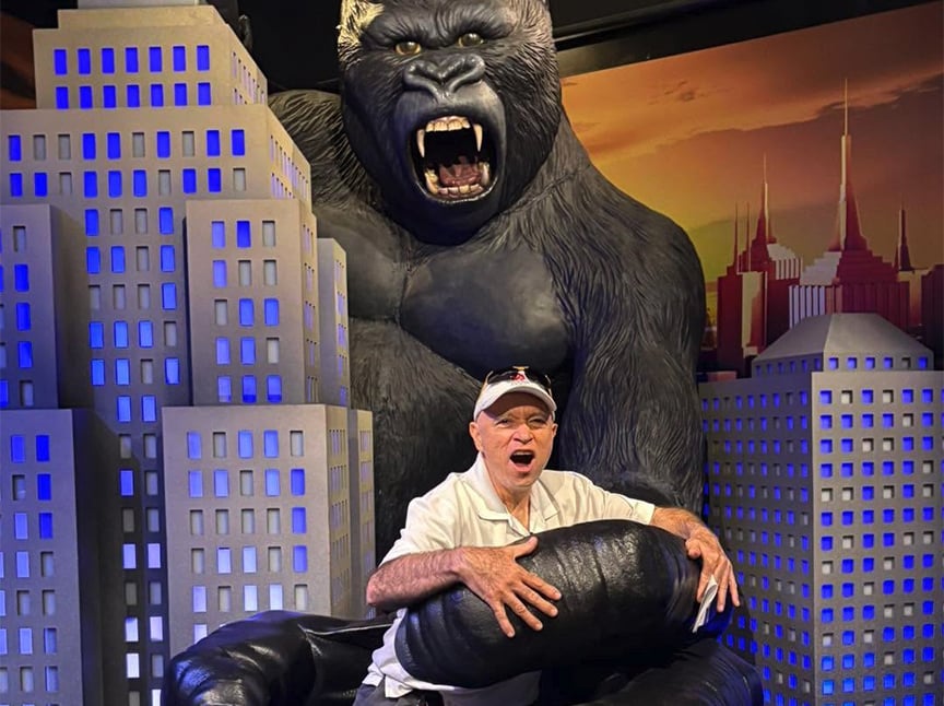 A man poses playfully in the giant hand of a King Kong statue, surrounded by model skyscrapers with a cityscape backdrop. He imitates a surprised expression, matching the roaring King Kong behind him.