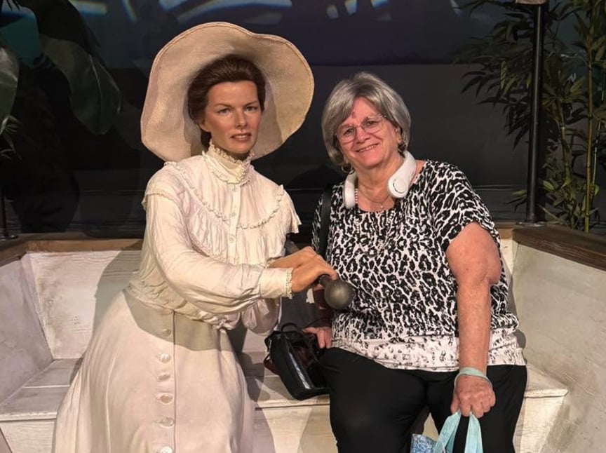 A woman with gray hair and glasses sits on a bench next to a wax figure dressed in a white Victorian-era dress and wide-brimmed hat. Both are smiling. The woman holds shopping bags and wears headphones around her neck.