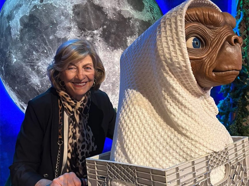 A smiling woman stands beside a life-sized E.T. model wrapped in a white blanket and sitting in a bicycle basket, with a large moon backdrop behind them.