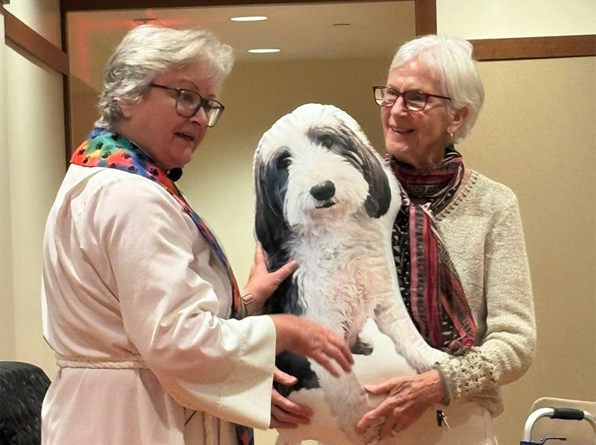 Two older women stand indoors holding a large, realistic pillow shaped like a fluffy dog. Both women are smiling and wearing glasses and sweaters, and the atmosphere appears friendly and warm.