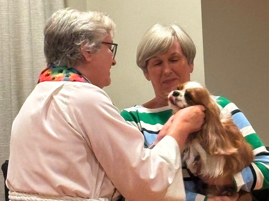 Two older women smile warmly as one gently pets a small, long-haired dog sitting on the others lap. One woman wears glasses and a colorful scarf, while the other wears a striped shirt and closes her eyes, enjoying the moment.