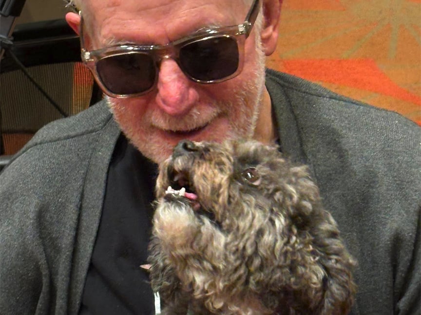 An older man wearing sunglasses and a gray sweater smiles as he sits closely with a small, curly-haired dog that looks up at him affectionately.