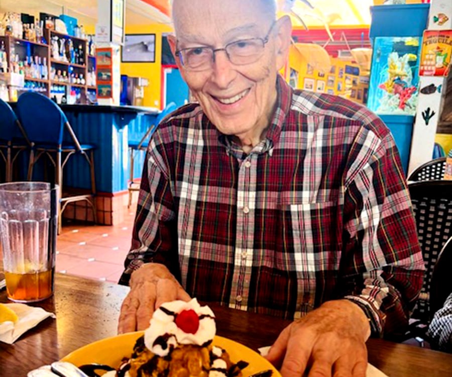 An elderly man wearing glasses and a plaid shirt smiles as he sits at a restaurant table with a dessert topped with whipped cream and a cherry in front of him. The colorful restaurant interior is visible in the background.