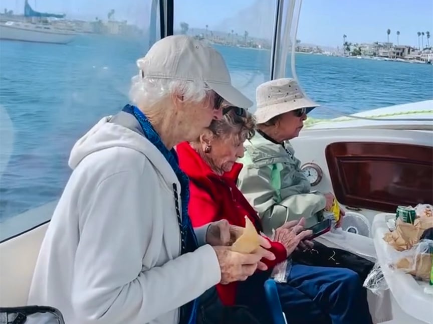 Three elderly women sit together on a boat, eating sandwiches and snacks. The boat is on a calm body of water with houses and palm trees visible in the background under a clear sky.