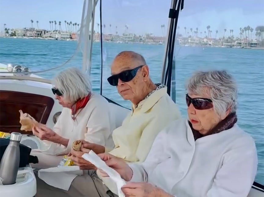 Three older adults wearing sunglasses sit on a boat, eating a meal together. Water and a coastal town with palm trees are visible in the background on a clear day.