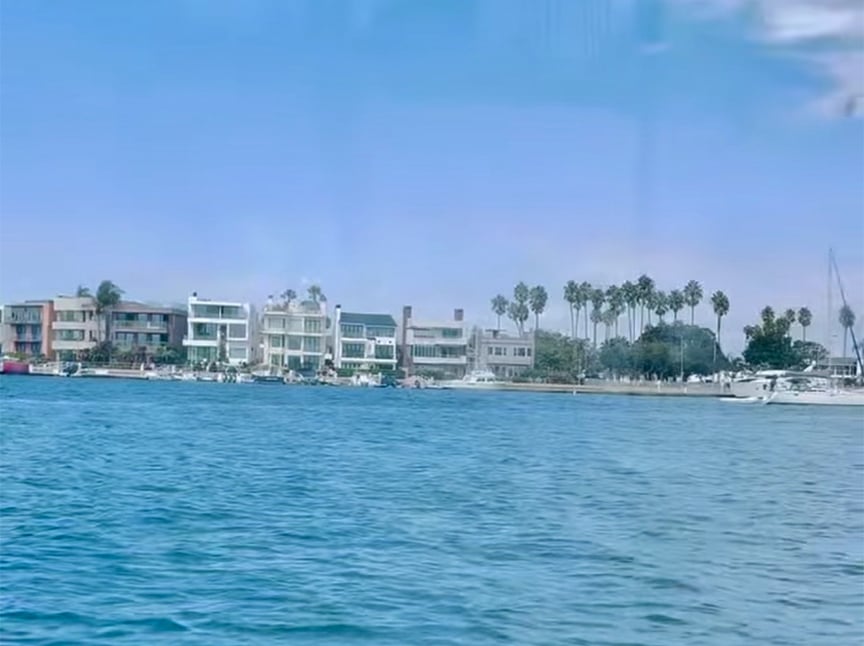 Waterfront view of modern houses with palm trees behind them, yachts and boats docked along the shore, and a blue sky above, seen from across a calm body of water.