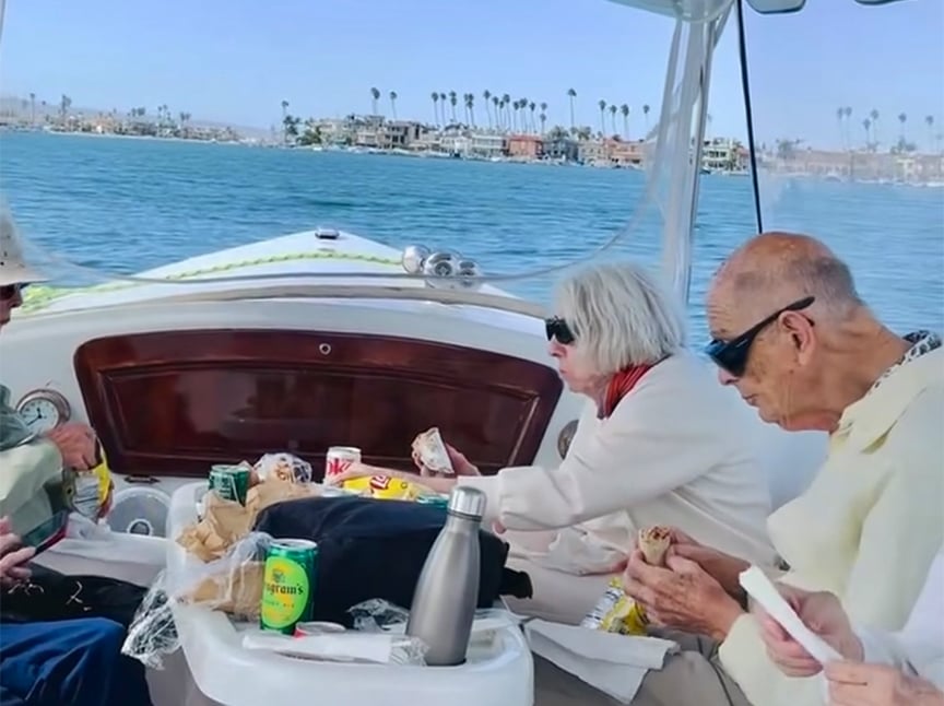 Three older adults wearing sunglasses eat sandwiches and snacks on a boat with water and a distant shoreline with palm trees in the background. Food, drinks, and a thermos are on the table in front of them.