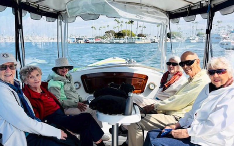 Six older adults are sitting and smiling on a boat with a marina and palm trees visible in the background on a sunny day.