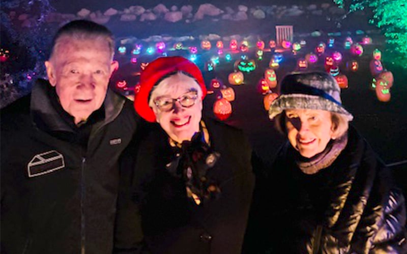 Three smiling older adults stand outdoors at night in front of a display of glowing, carved pumpkins. The people wear warm jackets and hats, and colorful lights illuminate the pumpkins in the background.