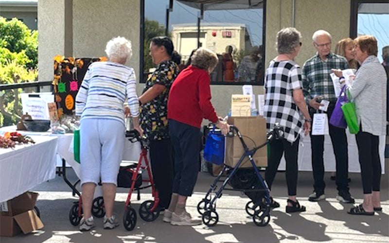 A group of older adults, some using walkers, gather and chat at an outdoor market stand displaying goods. The weather is sunny and the atmosphere appears friendly and social.
