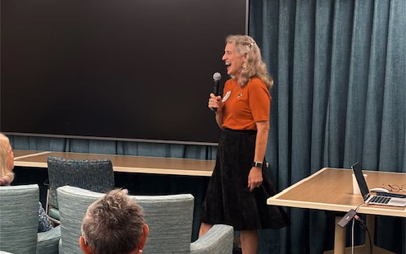 A woman with long gray hair holds a microphone and speaks to a seated audience in a room with blue curtains, a large screen, and a laptop on a table.