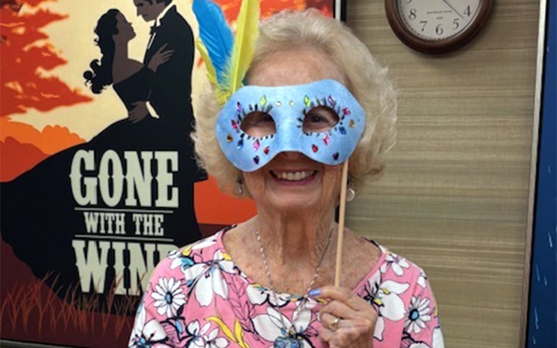 An older woman wearing a floral shirt smiles while holding a decorated blue mask with yellow and blue feathers in front of her face. Behind her is a Gone With the Wind movie poster and a clock on the wall.