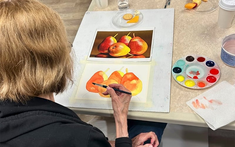 A person with light brown hair is painting three pears with watercolors, using a photo of pears as a reference. Paints, brushes, and a cup are on the table beside their painting.