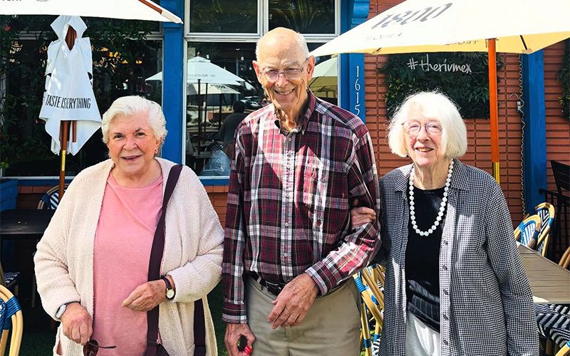 Three elderly people stand smiling outdoors at a café, with two women flanking a man in the center. They appear happy and are dressed in casual clothing; umbrellas and patio furniture are visible in the background.