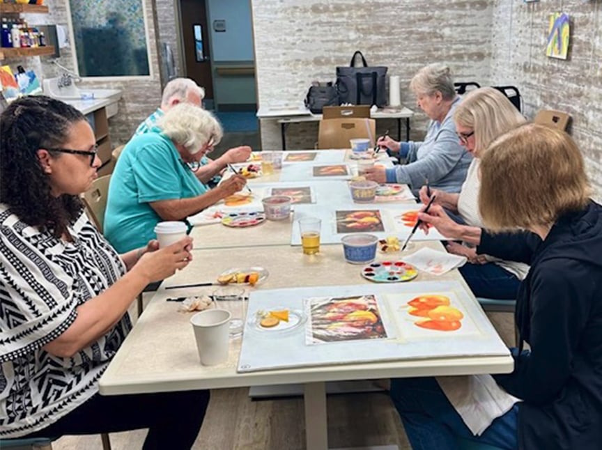 Six adults sit around a table in an art class, painting colorful fruit images on paper. Art supplies and drinks are on the table, and the setting is a bright, casual indoor room.