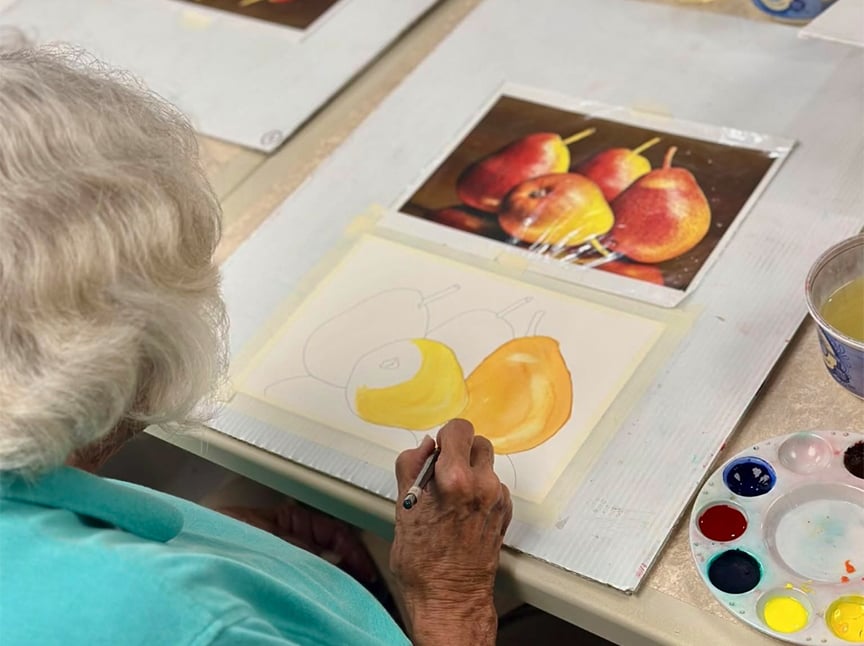 An elderly person with white hair paints a still life of pears, referencing a photo. The image shows the painting in progress, with colors being filled in, and a palette with paints nearby.