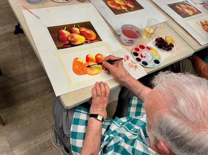 An elderly person in a green plaid shirt paints a watercolor picture of pears, referencing a photo of pears on the table. Art supplies, including brushes, paints, and water, are arranged nearby.